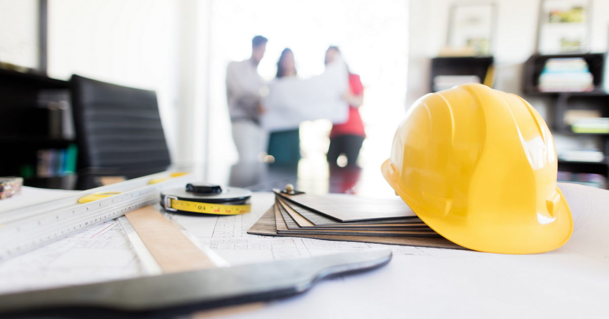 Desk with yellow helmet and other architectural equipment, ina distance behind three persons looking at a construction plan